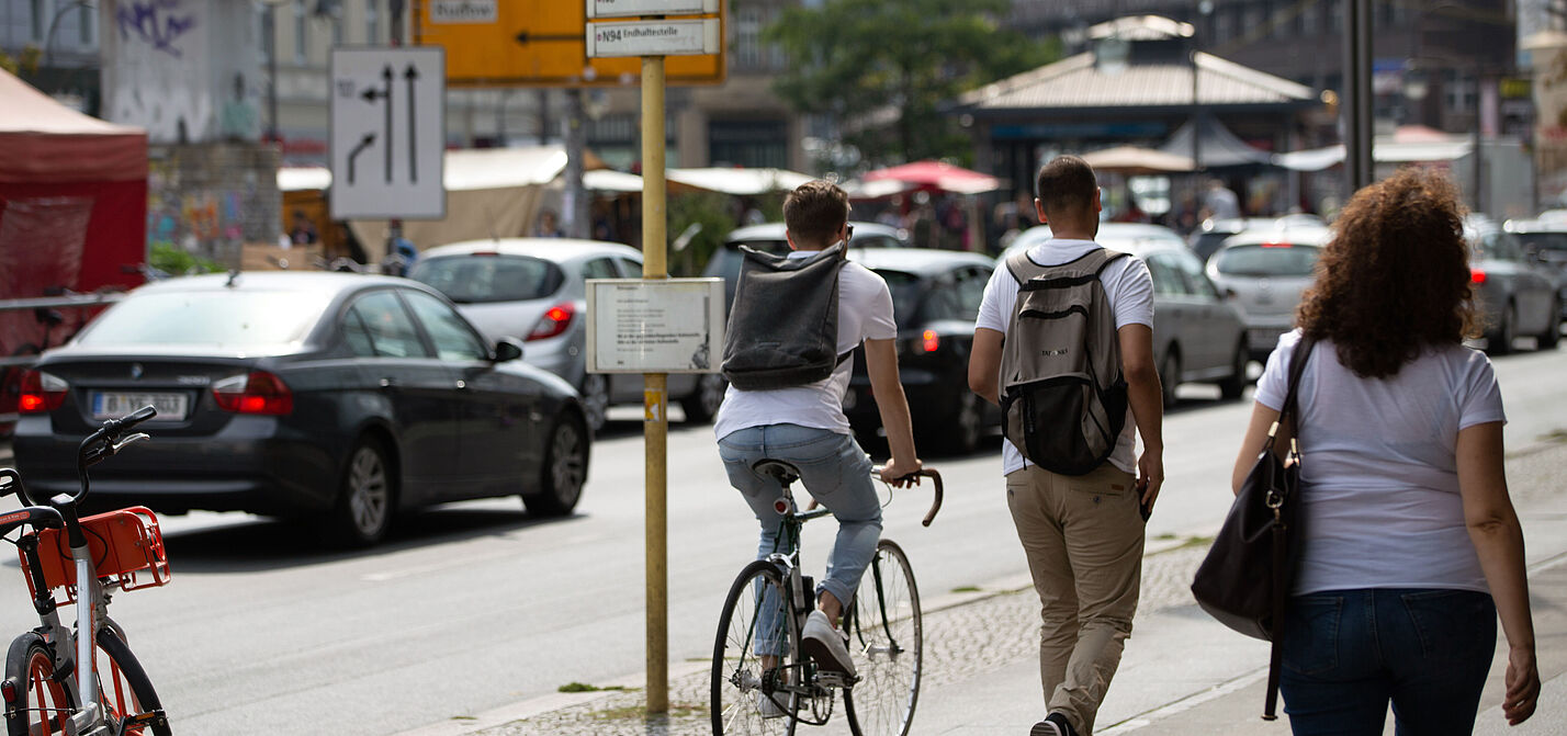 Eine Straßenszene: Ein Fahrradfahrer und zwei Fußgänger teilen sich einen gepflasterten Gehweg. Im Hintergrund parken Autos am Straßenrand.