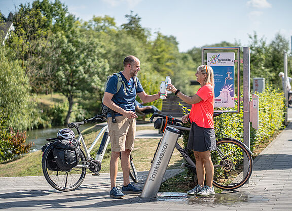 Zwei Radfahrende füllen Trinkflaschen an einem Trinkbrunnen des RuhrtalRadwegs auf.